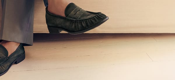 Ankle shot of a person wearing grey trousers and dark green suede Sam Edelman penny loafers.