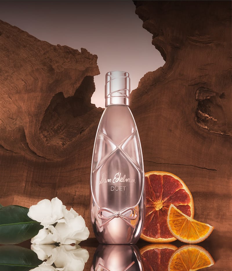 Sam Edelman Duet fragrance bottle displayed with white flowers and sliced oranges against a natural wood backdrop.