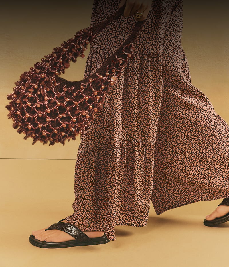 Brown woven bead shoulder bag carried by a model wearing a patterned outfit and black sandals.